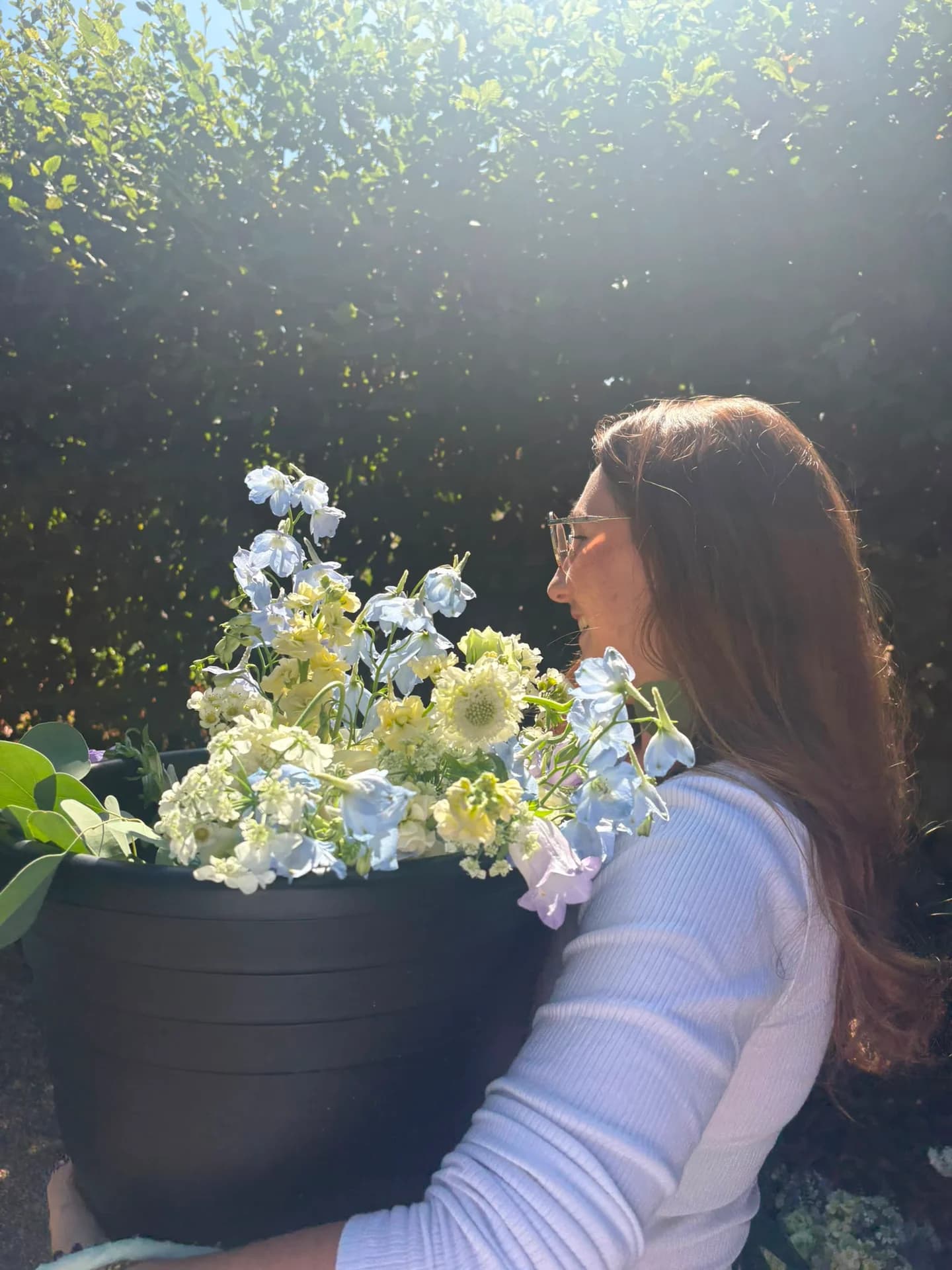 A woman carrying a bucket of pale blue, white and soft yellow flowers in the sunshine