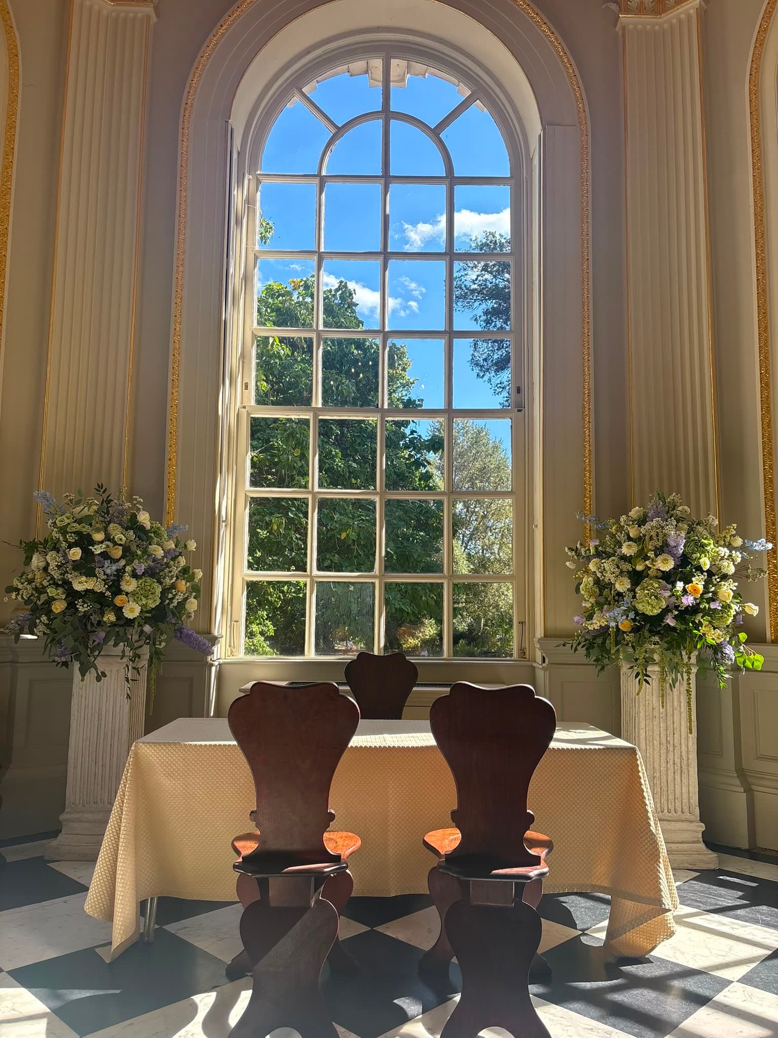 Two pastel floral arrangements on stone plinths framing a wedding ceremony table beneath an arched window